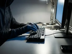 Vermummte Person tippt auf schwarzer Tastatur im dunklen Büro, beleuchtet von kaltem blauem Monitorlicht.