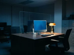 Einzelner Desktop-PC auf minimalistischem Schreibtisch, Glastrennwand reflektiert Monitorlicht, Büro in Stahl-Blau und Anthrazit mit warmem Schreibtischlampenlicht.