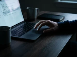 Hand zögert über Laptop-Trackpad, Bildschirm zeigt verdächtige E-Mail, dramatische Beleuchtung unterstreicht Phishing-Gefahr.